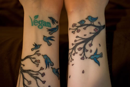 Tattoos of the word vegan in green ink and birds flying around some tree branches.