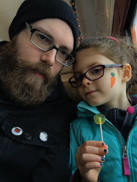 Michael bearded in a knit hat posing with his daughter who has a carrot painted on her check.
