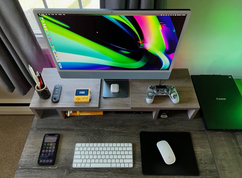 Overhead view of a blue 24-inch iMac, Playdate, Airpods, translucent Playstation 4 controller, keyboard, mouse, and Canon scanner arranged on a wooden desk.